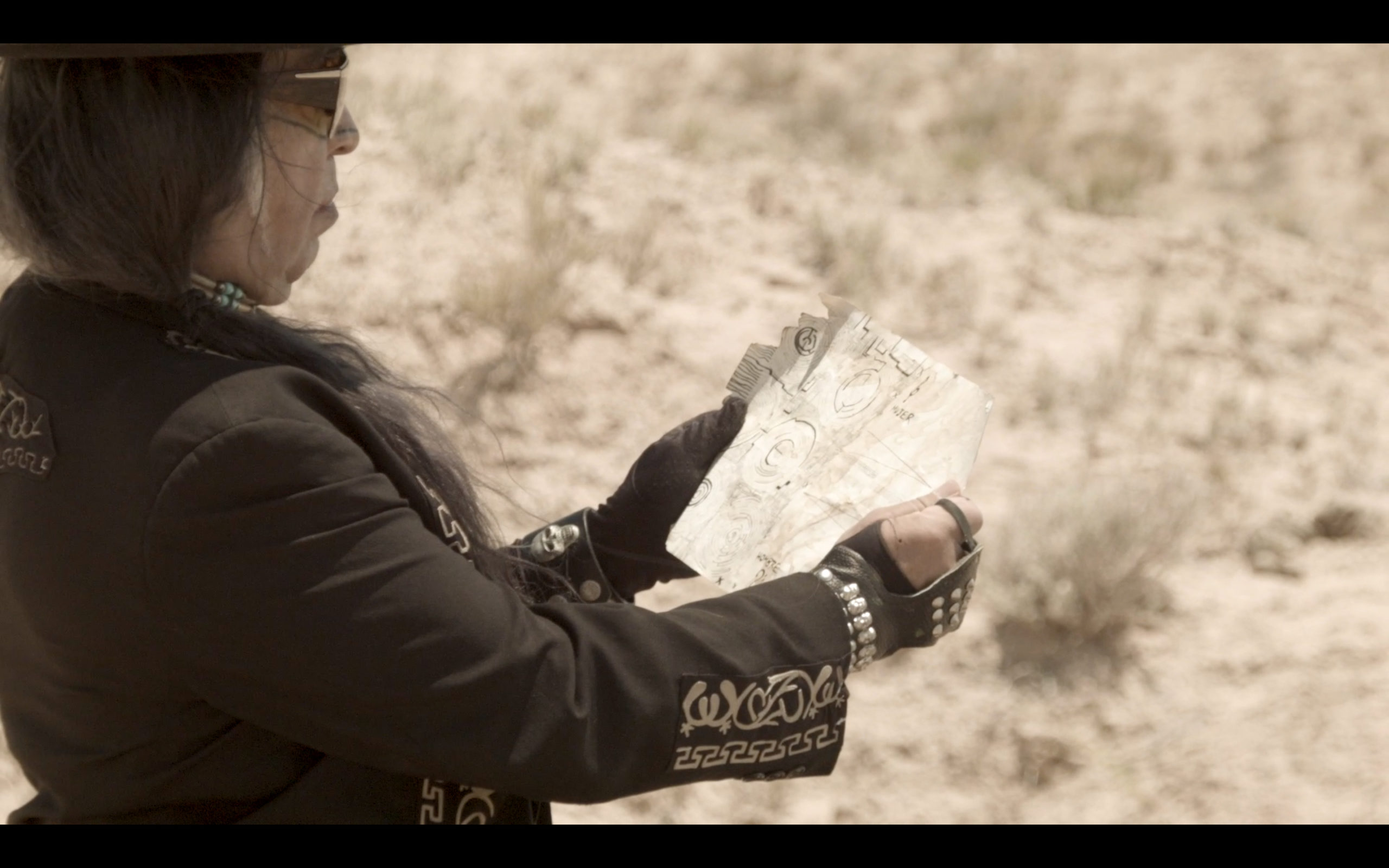 film still of a man holding a piece of paper (a map?) against a desert background